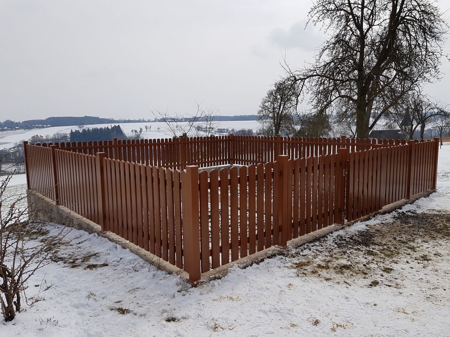 Ein Holzzaun im Schnee mit einem Hund, der neugierig davor steht und die winterliche Landschaft erkundet.